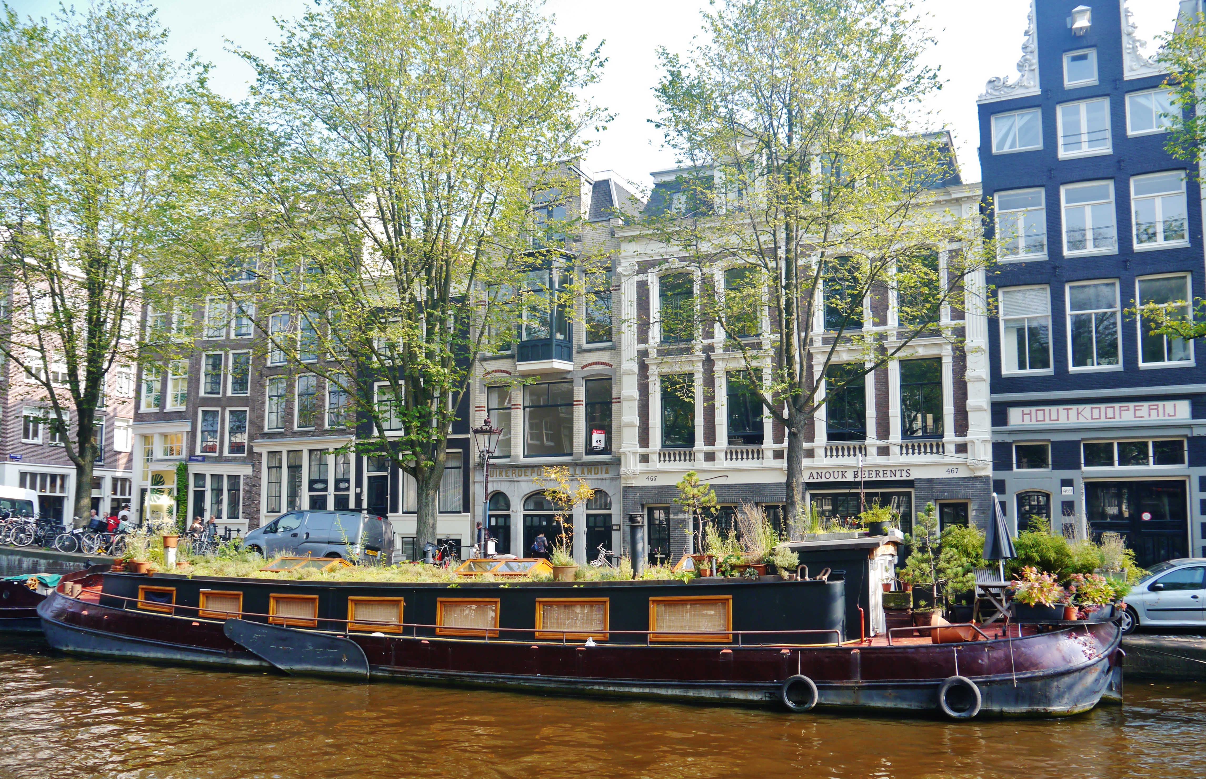 Lange woonboot met houten panelen en groen aan een gracht, op de achtergrond de historische gebouwen en bomen van Amsterdam