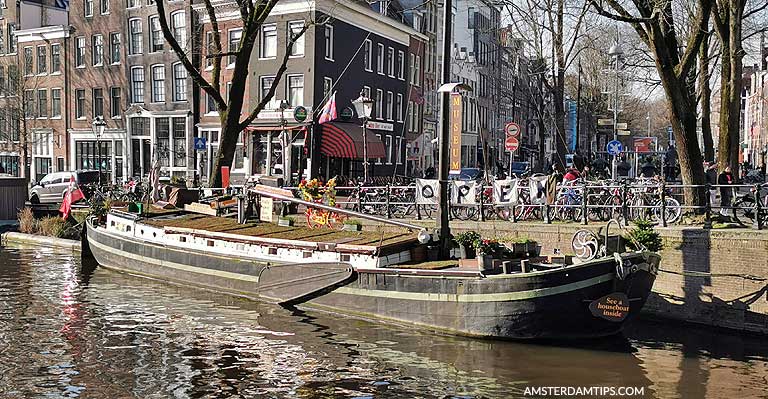 Woonboot aangemeerd langs een gracht met bakstenen gebouwen en voetgangers aan de kade in Amsterdam
