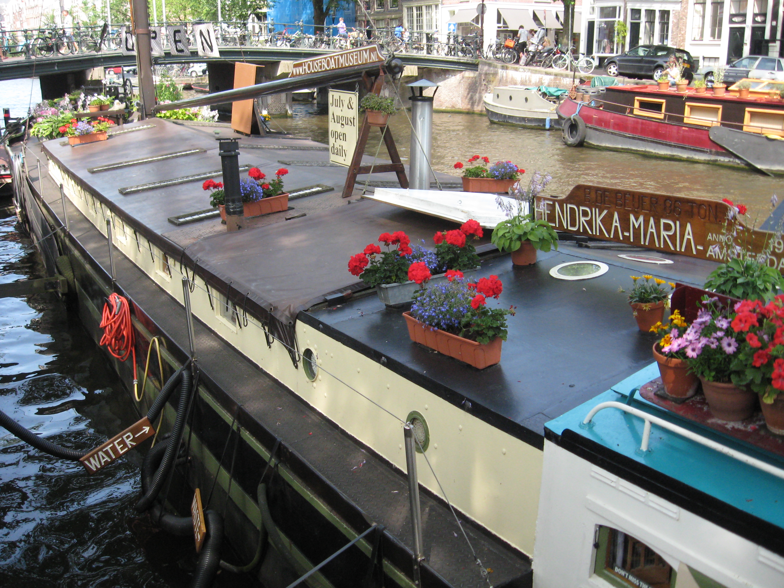 Woonboot genaamd Hendrika-Maria aan Amsterdamse gracht, gedecoreerd met rode en paarse bloemen op het dak, met brug en andere boten zichtbaar op de achtergrond