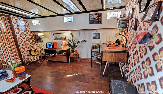 Gemeubileerd interieur van een woonboot met houten vloeren, muren met retro-patroon, een kleine tv, stoelen en planten.