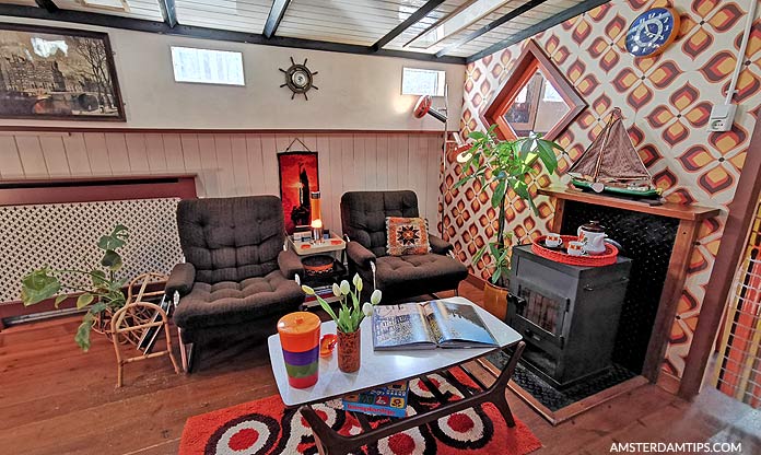 Cozy canal houseboat interior with two armchairs, coffee table, small stove, patterned wallpaper, and nautical decor including a ship's wheel and model boat.