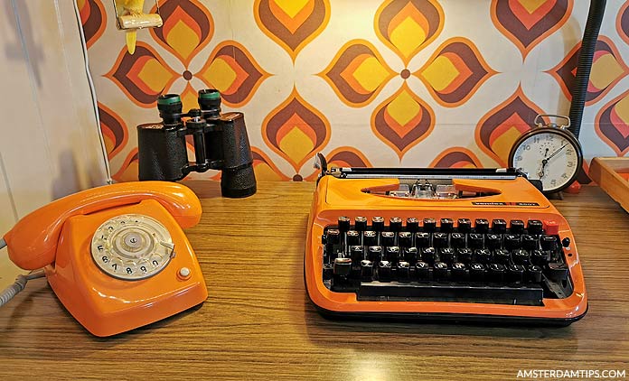 Oranje draaistelefoon, typemachine, verrekijker en klok op een houten tafel tegen een achtergrond van retro-patroon behang.