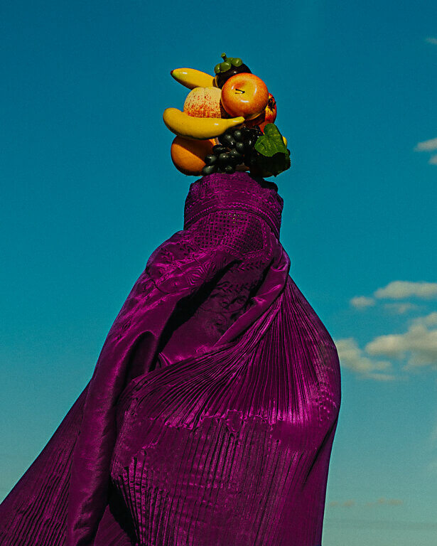 A figure draped in a rich purple garment holds a vibrant arrangement of fruits including bananas, oranges, and grapes against a clear blue sky