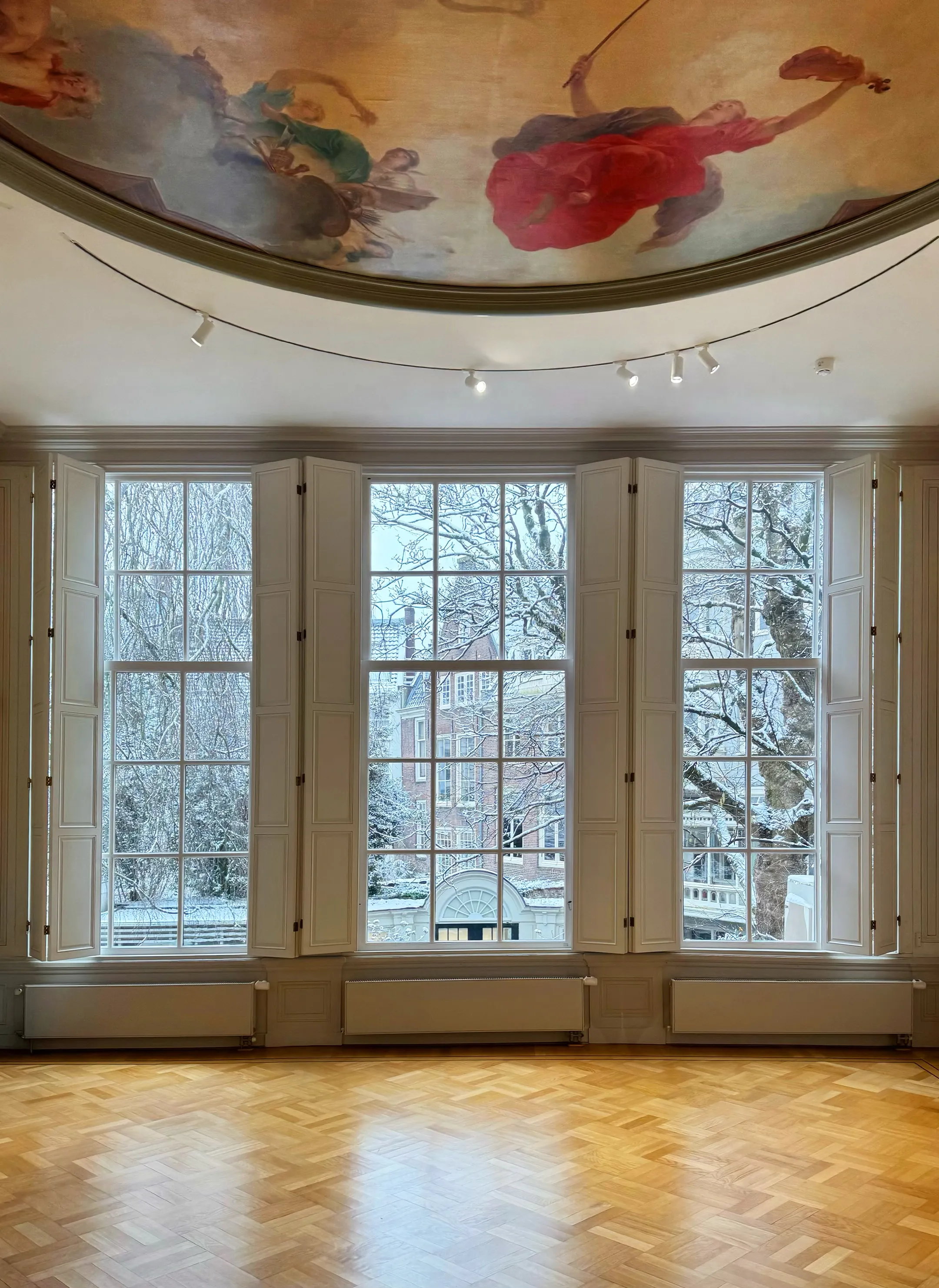 Interior of a gallery room with a painted ceiling fresco, large windows, and wooden floor