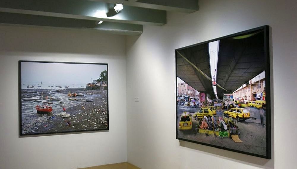 Two framed color photographs on a white gallery wall: left shows a beach with boats, right depicts a street scene with yellow taxis.