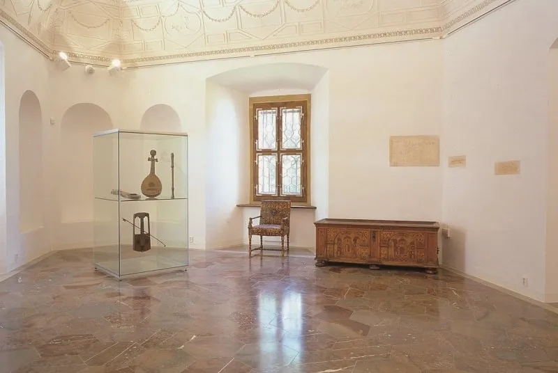 Interieur van een museumgalerij met een glazen vitrine, een houten kist en een stoel bij een raam
