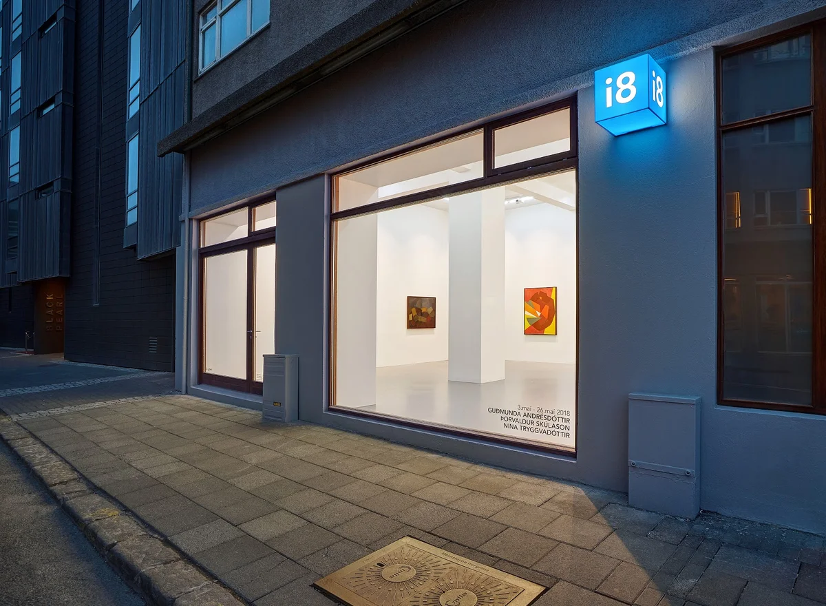 Buitenkant van i8 Gallery in de schemering met verlicht blauw i8-bord en grote glazen ramen met kunstwerken aan de binnenkant