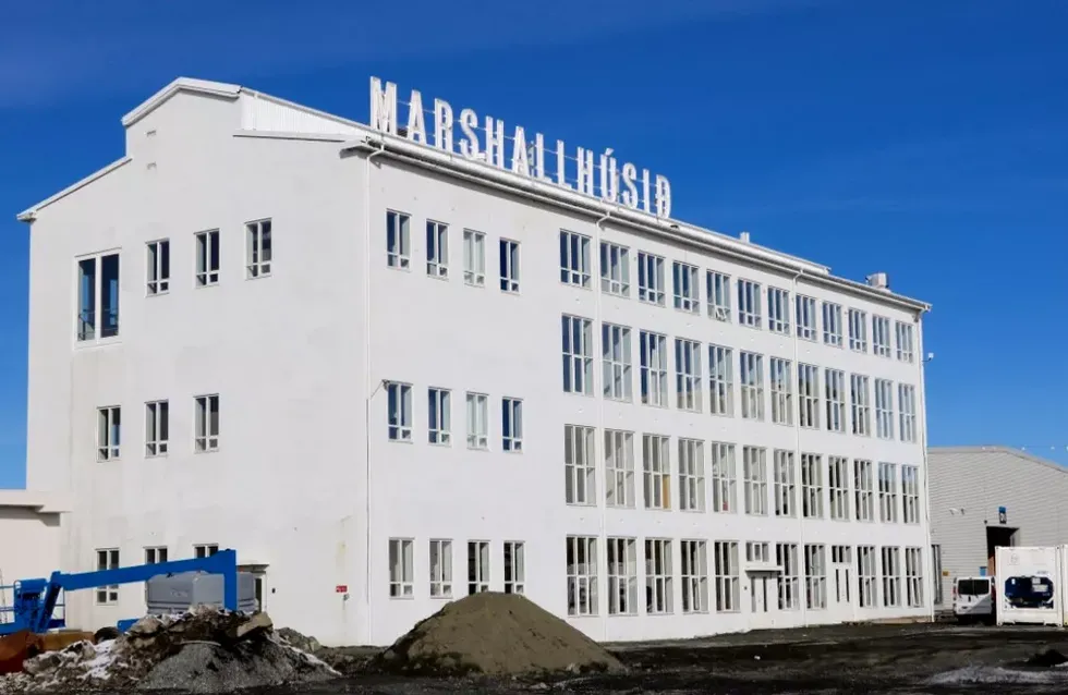 Gran edificio blanco de varios pisos con el letrero MARSHALLHÚSIN en el techo contra un cielo azul despejado, con materiales de construcción y una grúa azul en primer plano.