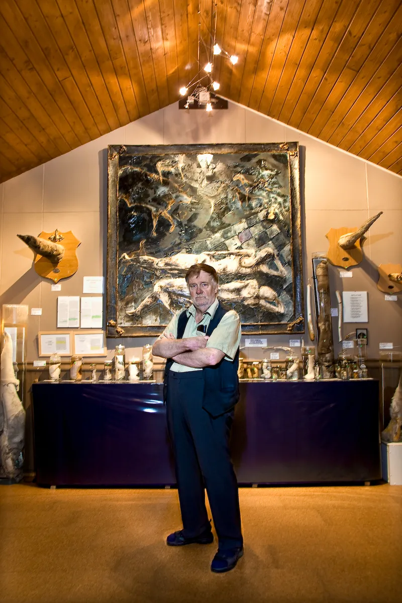 Man standing with arms crossed in front of display cases filled with specimens, with a large framed painting and mounted horn-like objects on the walls inside a museum with a wooden ceiling