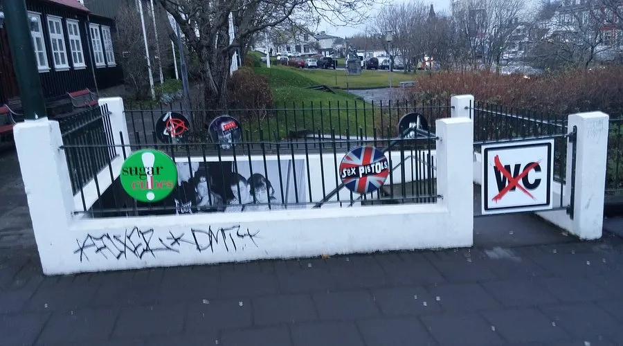 Cerca de hormigón blanco con verjas de metal negro cubiertas de pegatinas punk y grafitis, con un letrero de WC tachado en rojo a la derecha