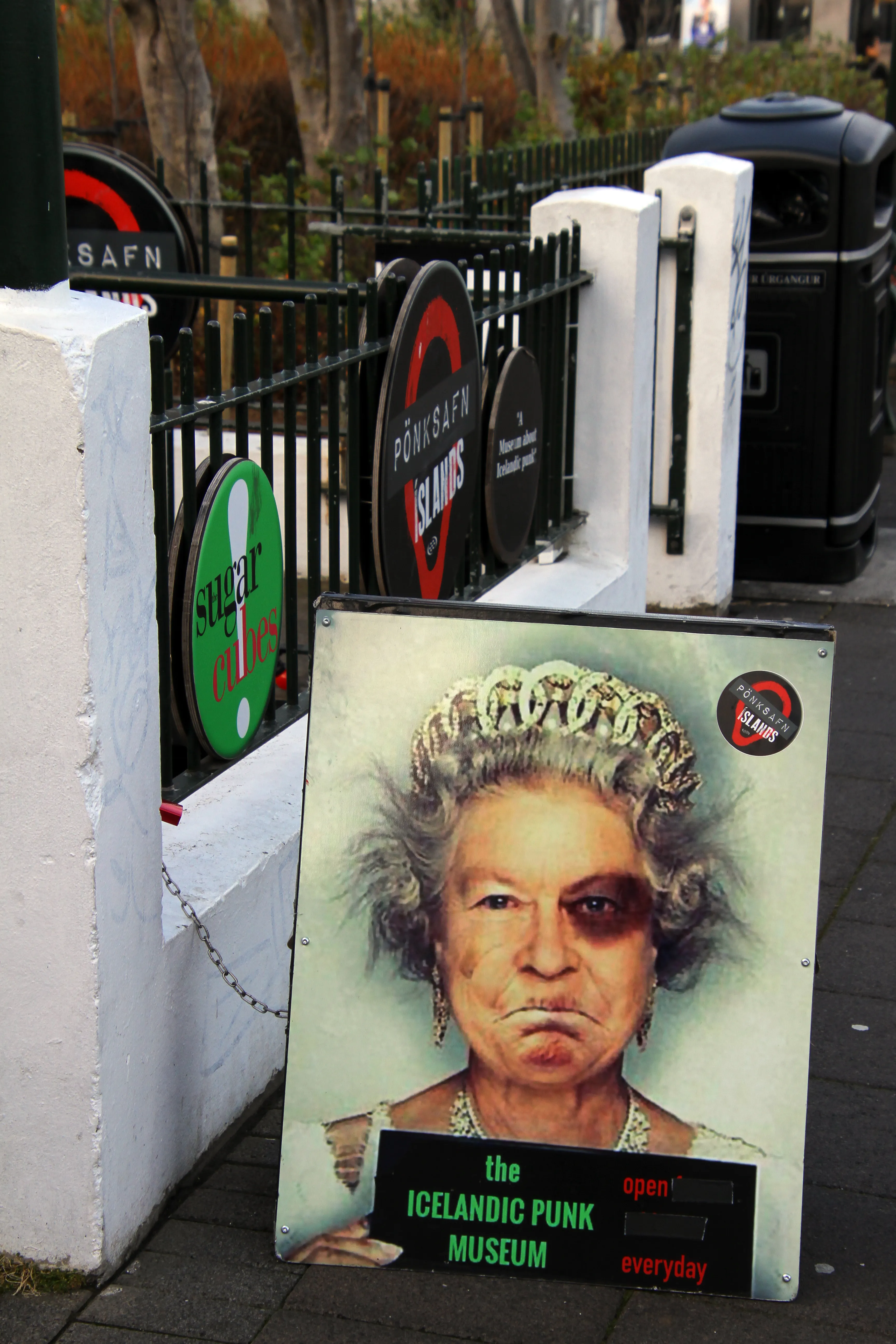 Caballete publicitario para el Icelandic Punk Museum con un retrato al estilo punk de la Reina Isabel II, con carteles circulares del local sobre una valla de metal negro detrás.