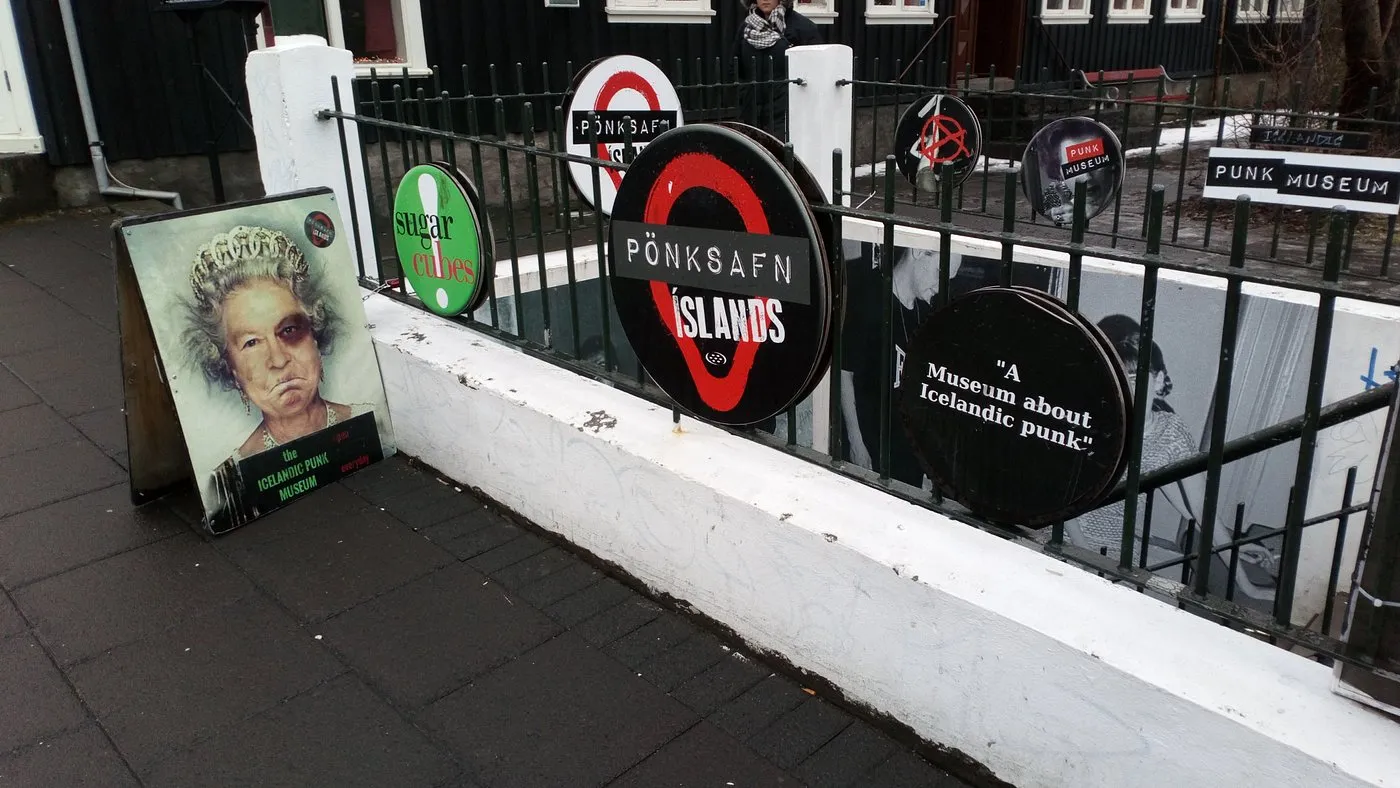 Vista exterior de la entrada del Icelandic Punk Museum, que cuenta con una valla de metal negro adornada con letreros circulares que se asemejan al logotipo del metro de Londres, incluido uno que dice "PUNKSAFN ÍSLANDS", y un cartel publicitario de caballete en la acera.