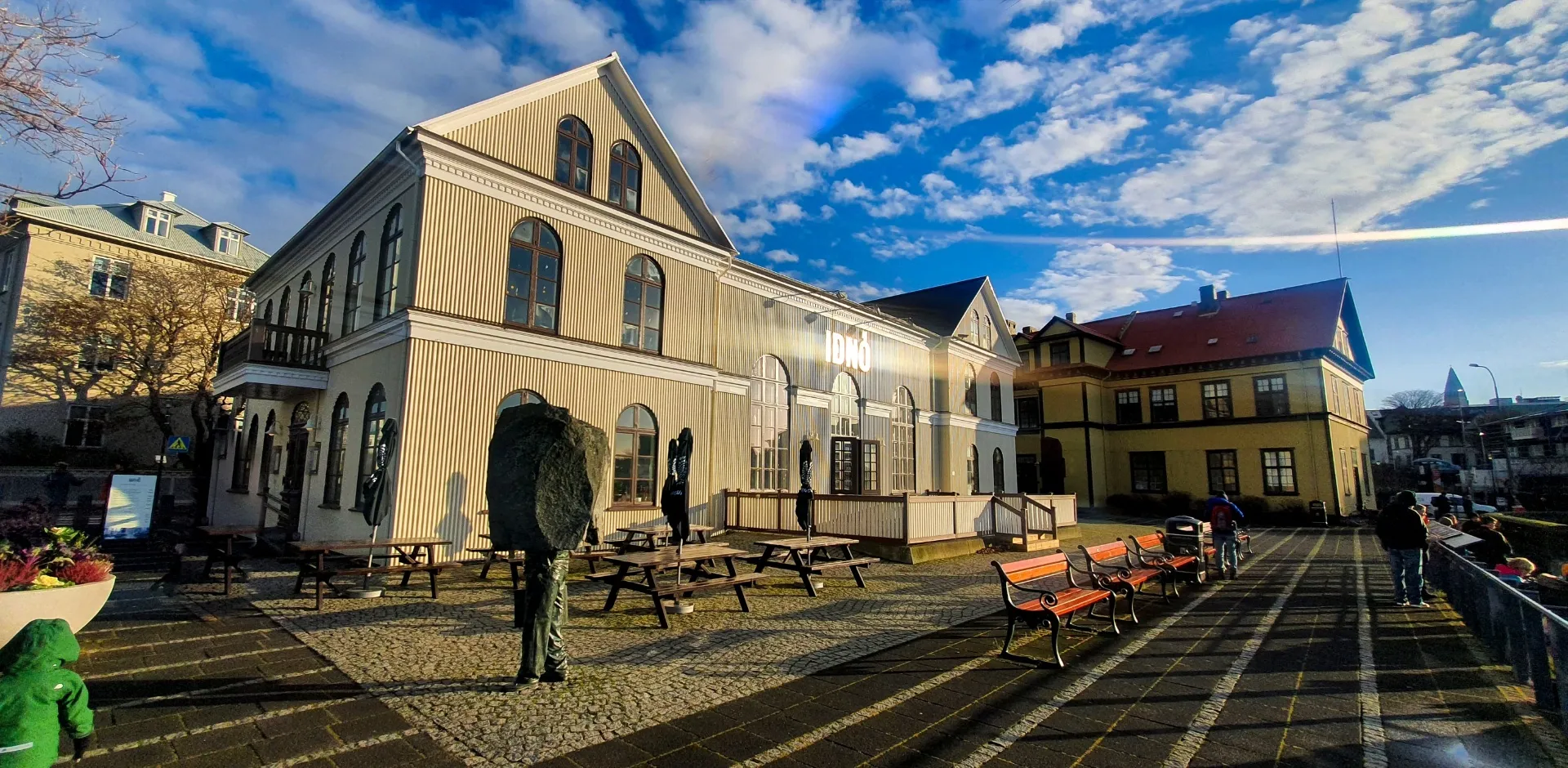 Historic wooden theatre building with cream-colored vertical siding, arched windows, and gabled roof, set against a blue sky with a paved plaza and benches in the foreground