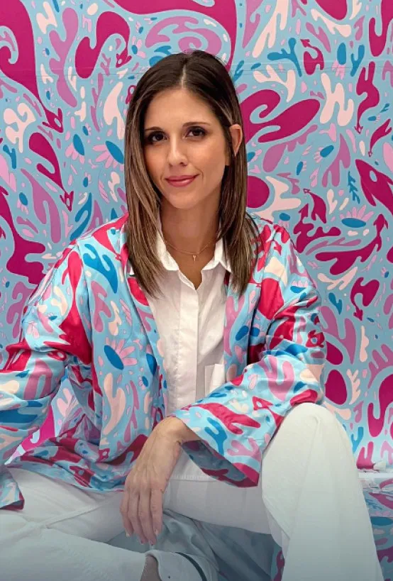 A woman in a colorful patterned jacket sitting in front of a blue and pink abstract wall