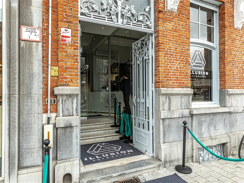 Entrada de fachada de ladrillo del museo Illusion Antwerpen con una ornamentada puerta de metal blanco, felpudo de la marca y logotipo visible en el escaparate