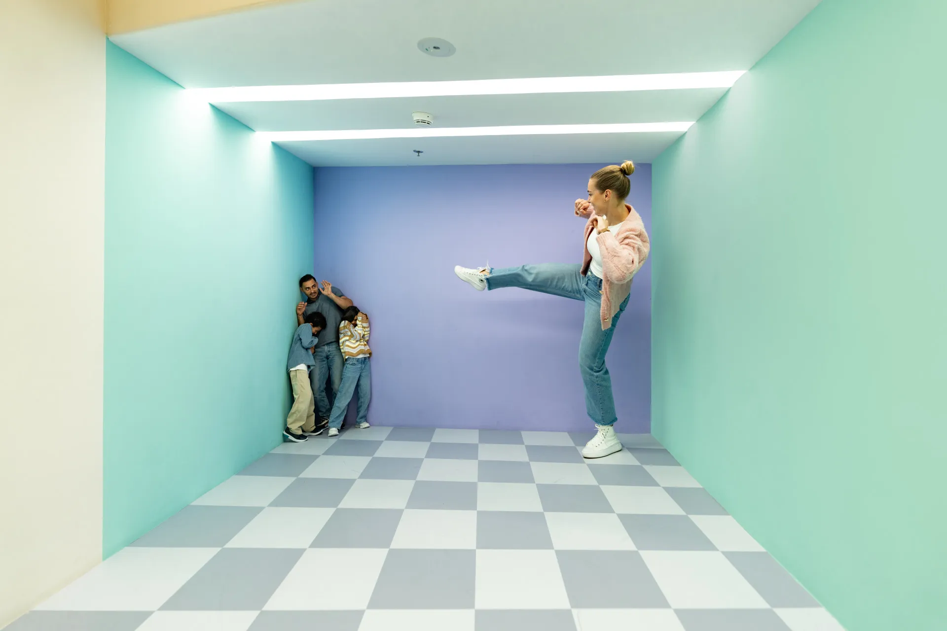 Personas posando dentro de una habitación de ilusión óptica con paredes de color verde menta, una pared trasera de color púrpura y un suelo a cuadros grises y blancos, creando un efecto de perspectiva forzada donde una mujer parece gigante mientras un grupo se ve diminuto en la esquina.