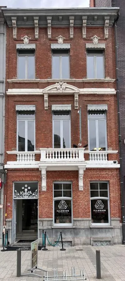 Fachada de ladrillo rojo con ornamentos de piedra blanca, un balcón con balaustrada y ventanas en la planta baja que muestran el logotipo de Illusion Antwerpen