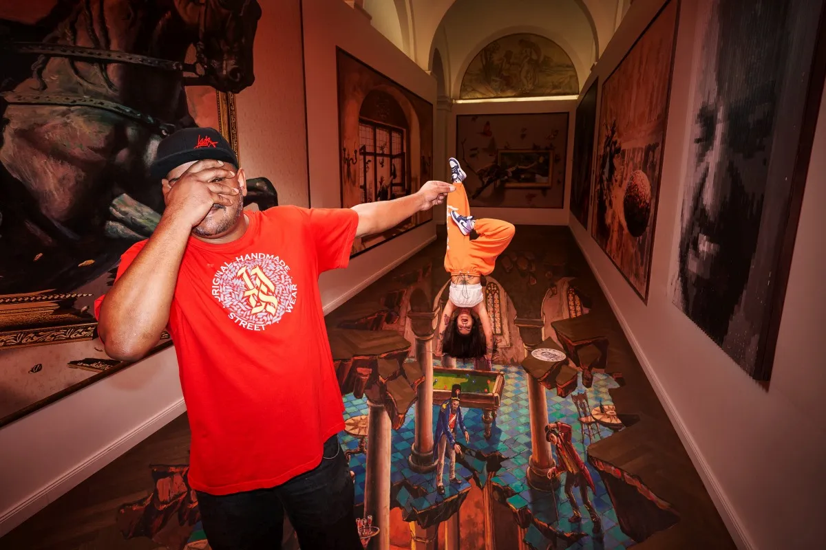 Een man in een rood shirt staat in een galerie en bekijkt een 3D-illusieschilderij op de vloer, met grote portretten aan de muren.