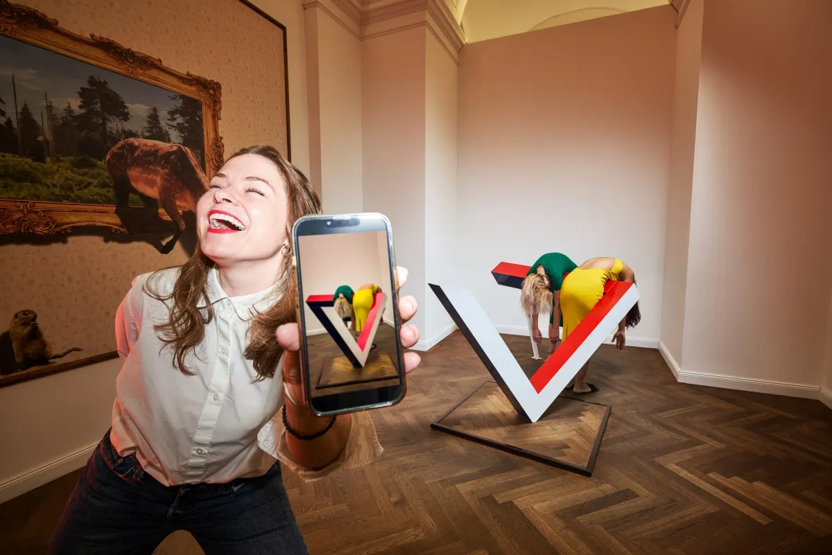 Een lachende vrouw die een smartphone vasthoudt voor een 3D-optische illusiesculptuur in een museumgalerij.