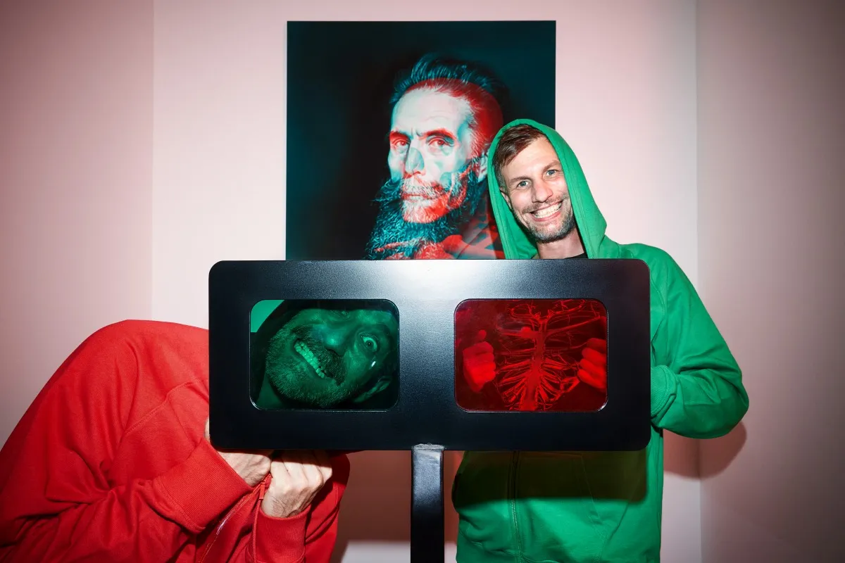 Twee mensen in kostuums met capuchons staan naast een 3D-illusie kunst display met rode en groene lenzen en een portret van een man met een baard.