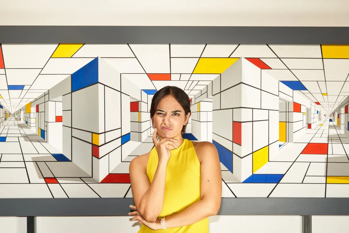 Een vrouw in een geel topje staat voor een kleurrijke geometrische optische illusie muurschildering met blauwe, rode, gele en witte kubussen die een 3D-effect creëren.