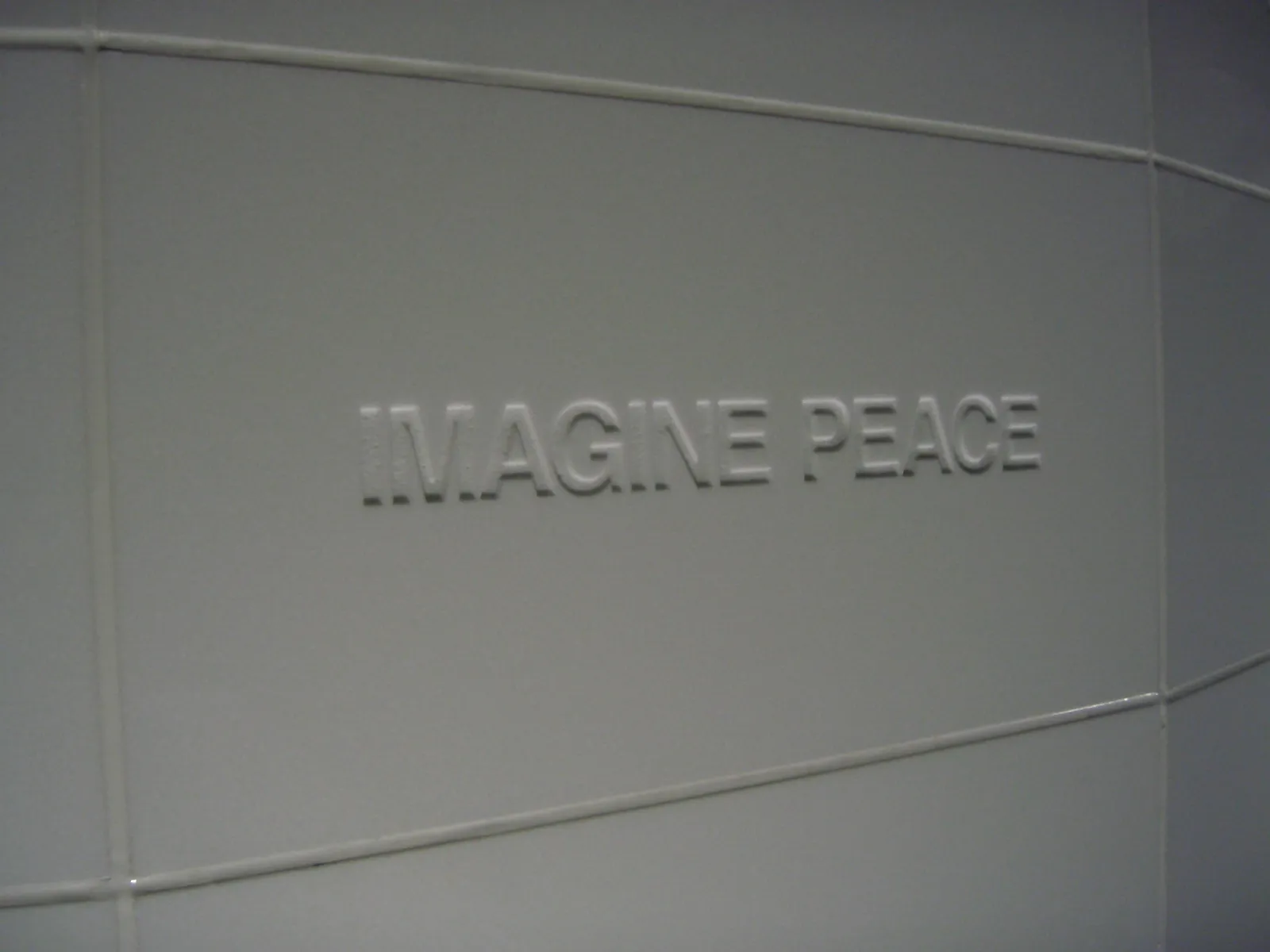 Wytłoczony napis "IMAGINE PEACE" na zakrzywionym szarym panelu z cienkimi poziomymi liniami biegnącymi przez powierzchnię