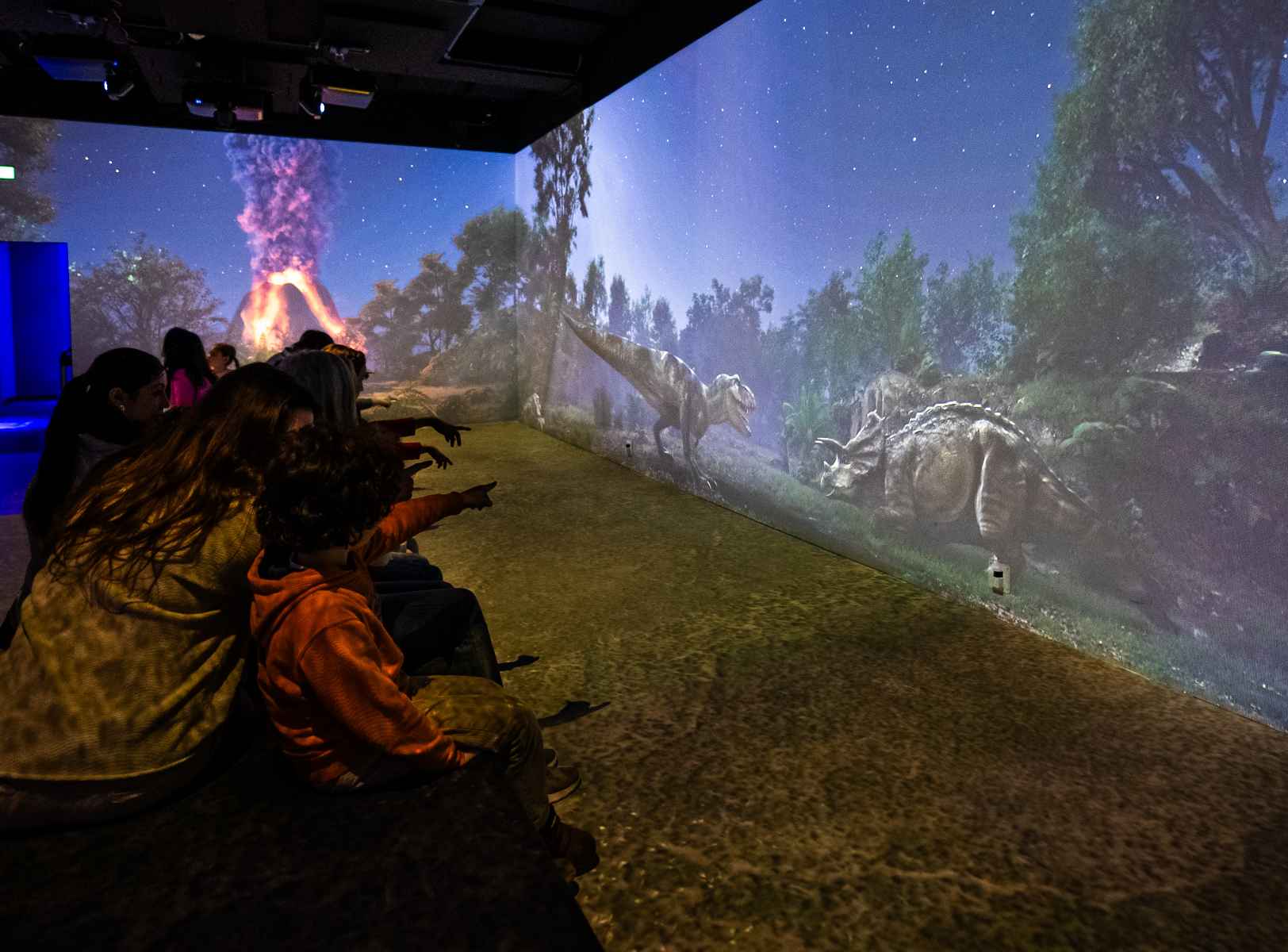 Bezoekers zitten in een projectieruimte van Immersium Vienna, omringd door een panoramisch prehistorisch landschap met dinosaurussen en een uitbarstende vulkaan.