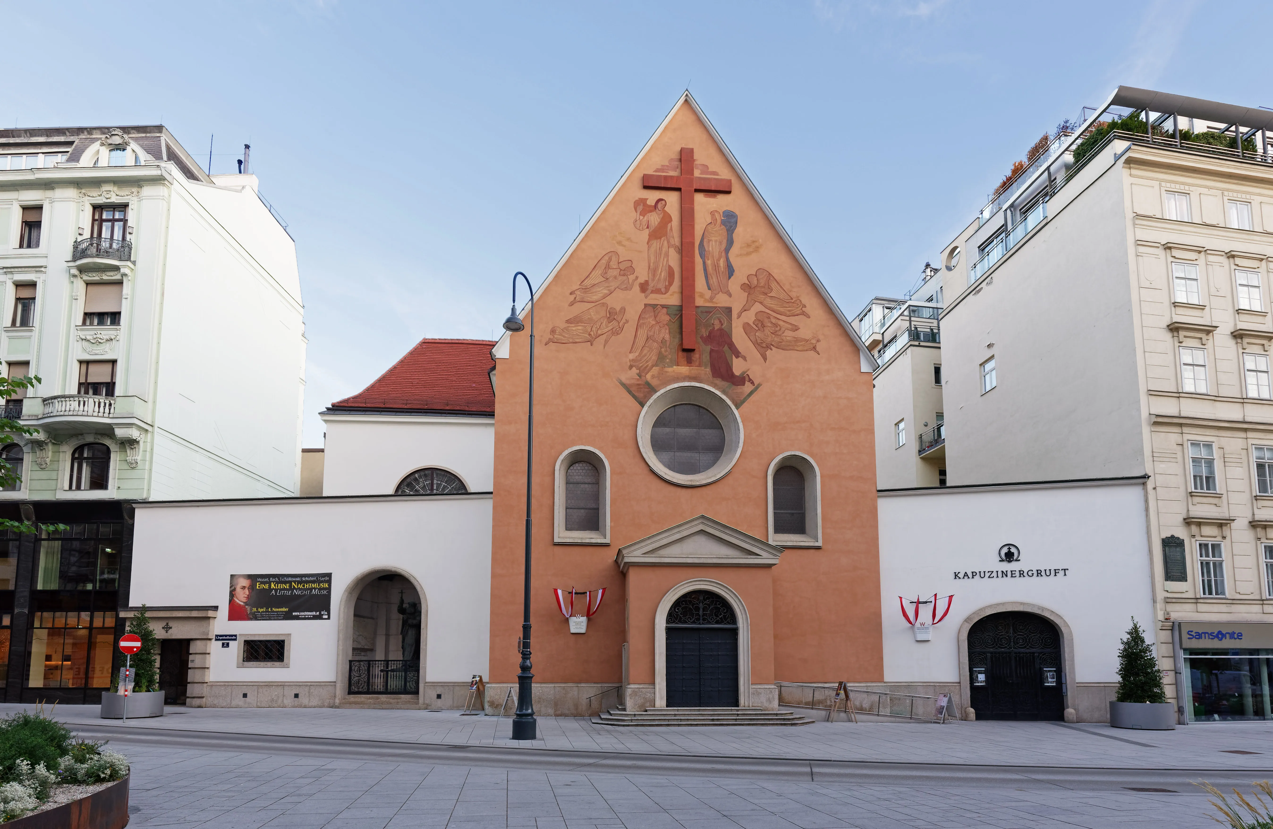 Kościół Kapucynów z pomarańczową fasadą, dużym krzyżem i religijnymi dziełami sztuki nad wejściem, otoczony budynkami miejskimi.