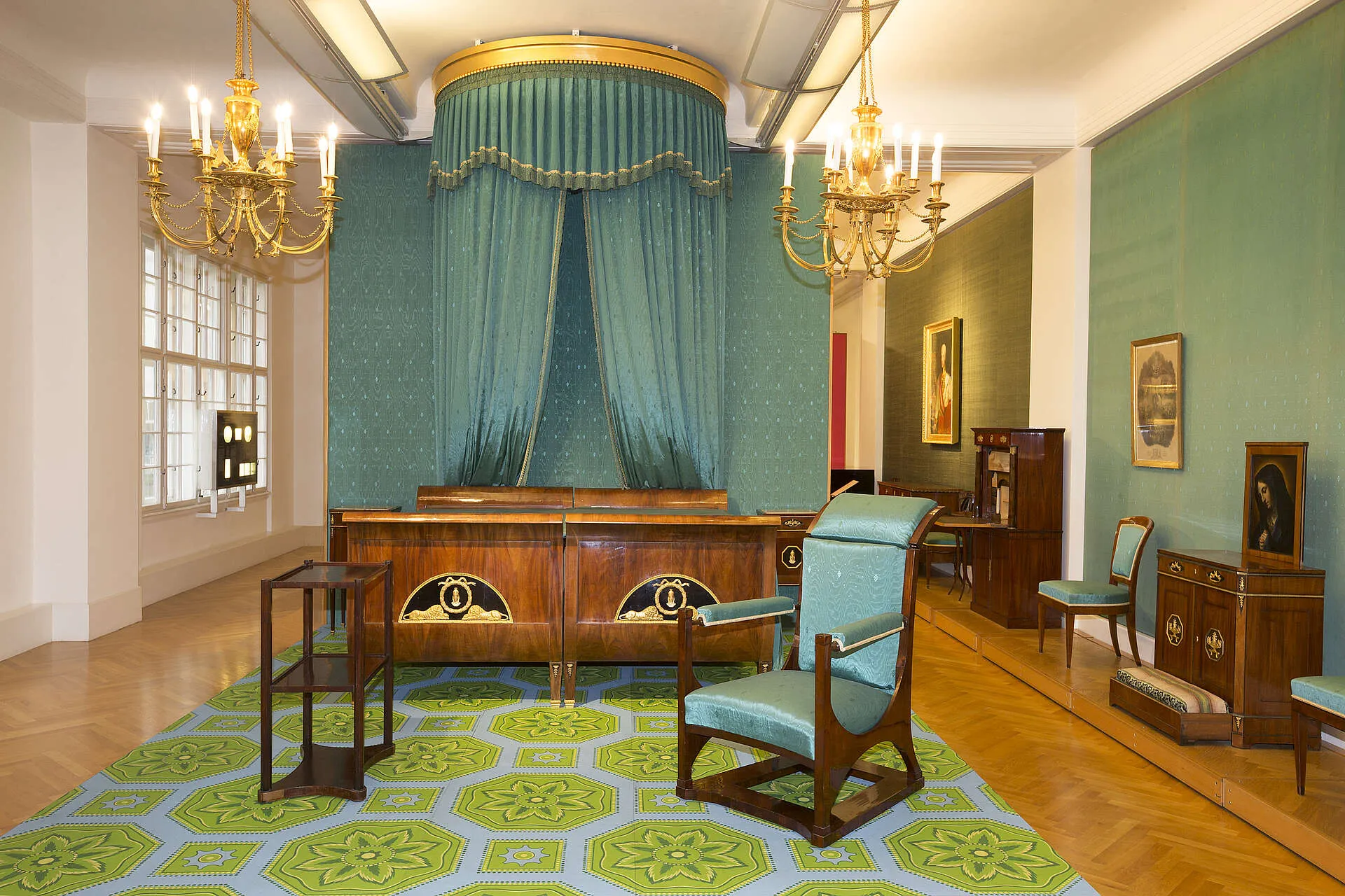 Habitación ornamentada con paredes de color verde azulado, una gran cama con dosel drapeado, candelabros y muebles de época que incluyen un escritorio y sillas de madera.