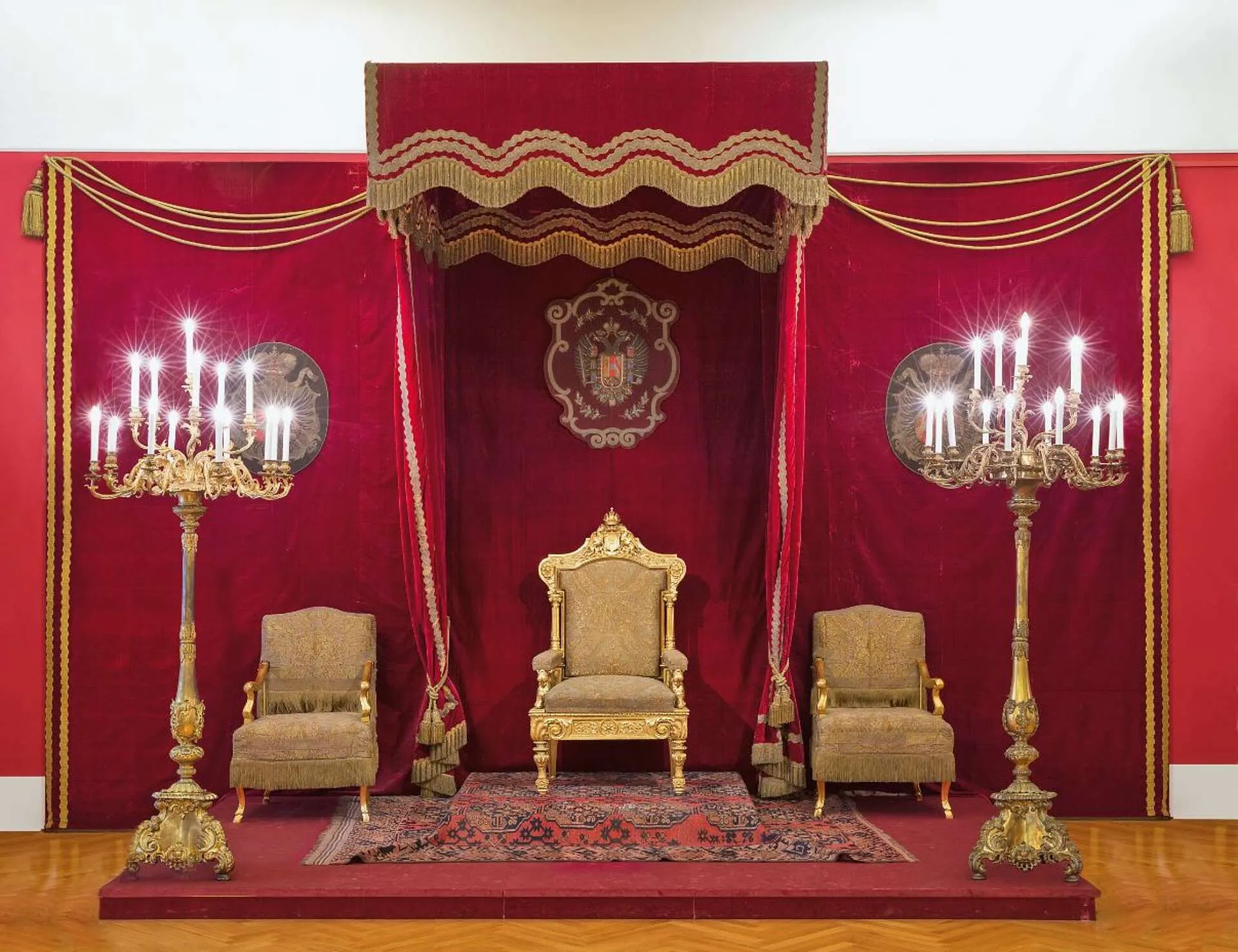 Un trono dorado con dos sillones bajo un dosel de terciopelo rojo, flanqueado por altos candelabros y un emblema heráldico sobre una pared roja.