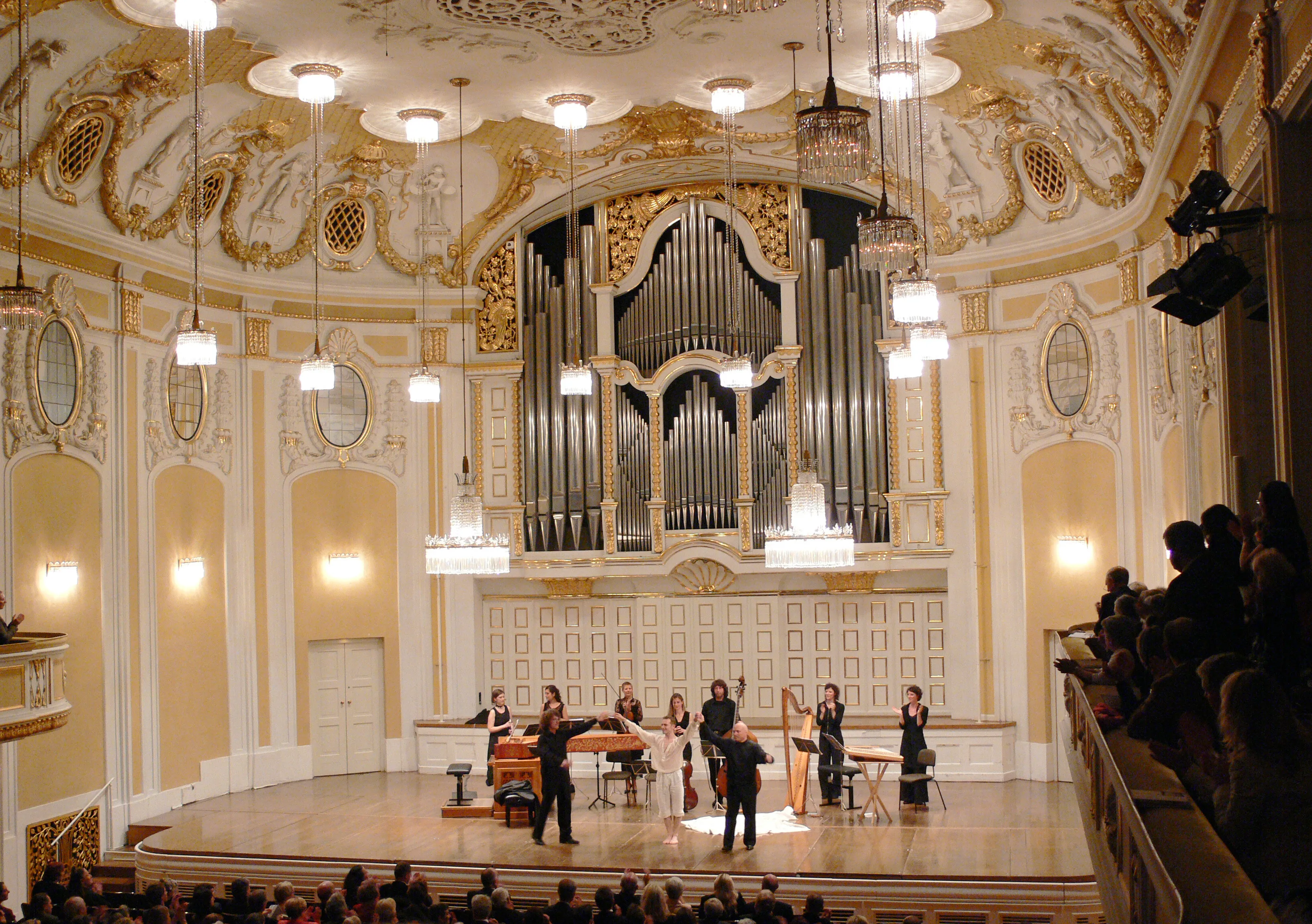 Ornate baroque concert hall interior with a large pipe organ on the back wall, musicians performing on stage, and an audience seated in the foreground