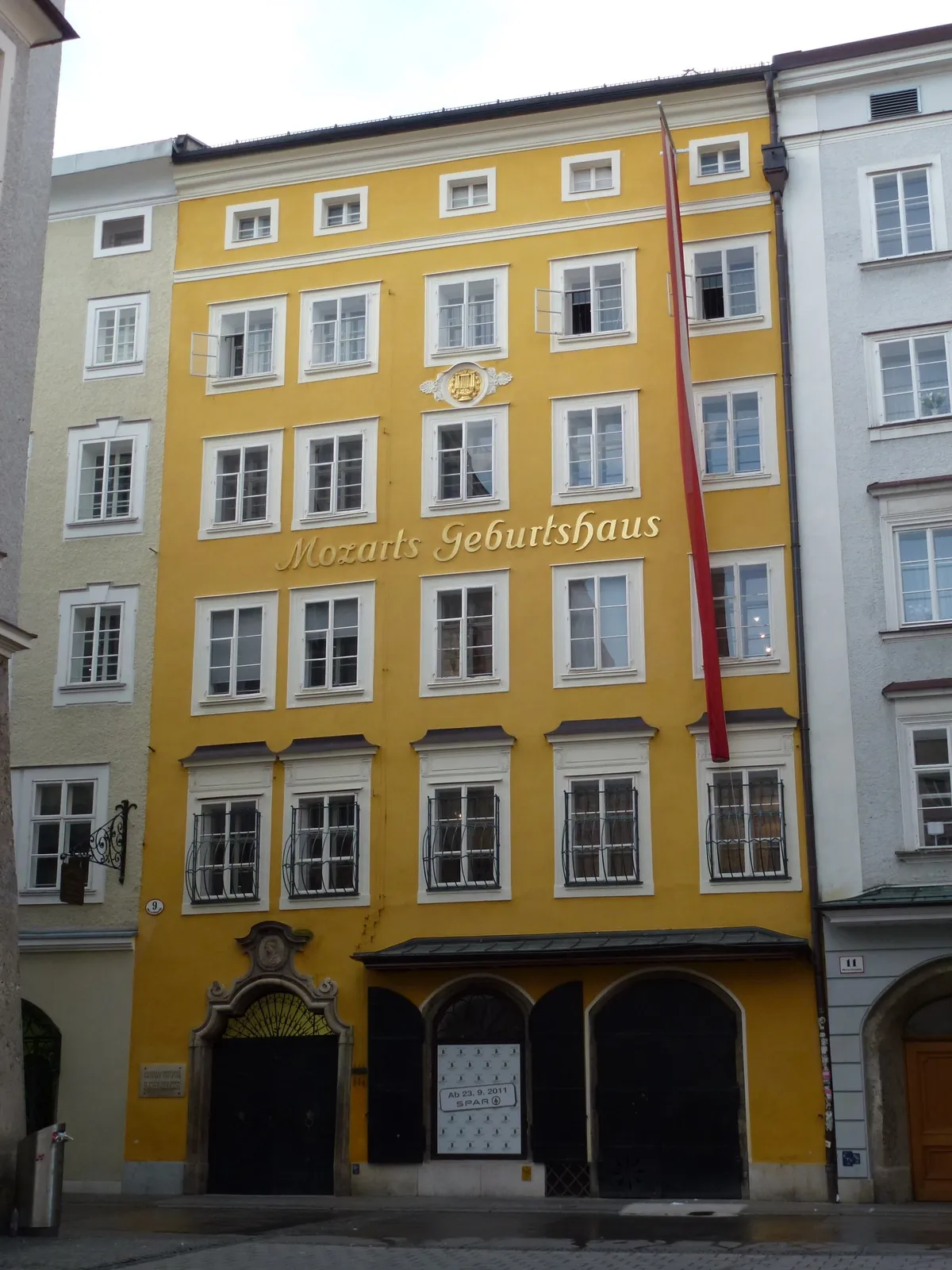 Yellow multi-story building facade with white-framed windows and gold lettering reading 'Mozarts Geburtshaus', with a red banner hanging from the upper floors