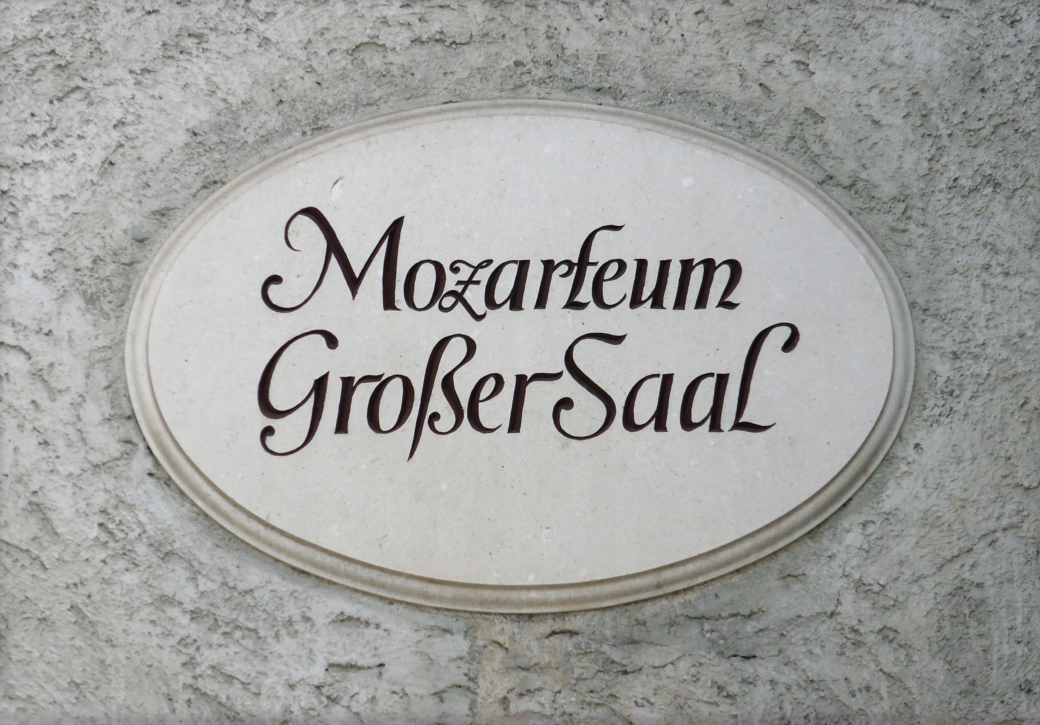 Oval white plaque with black Fraktur-style text reading "Mozarteum Großer Saal" mounted on a textured gray stone wall