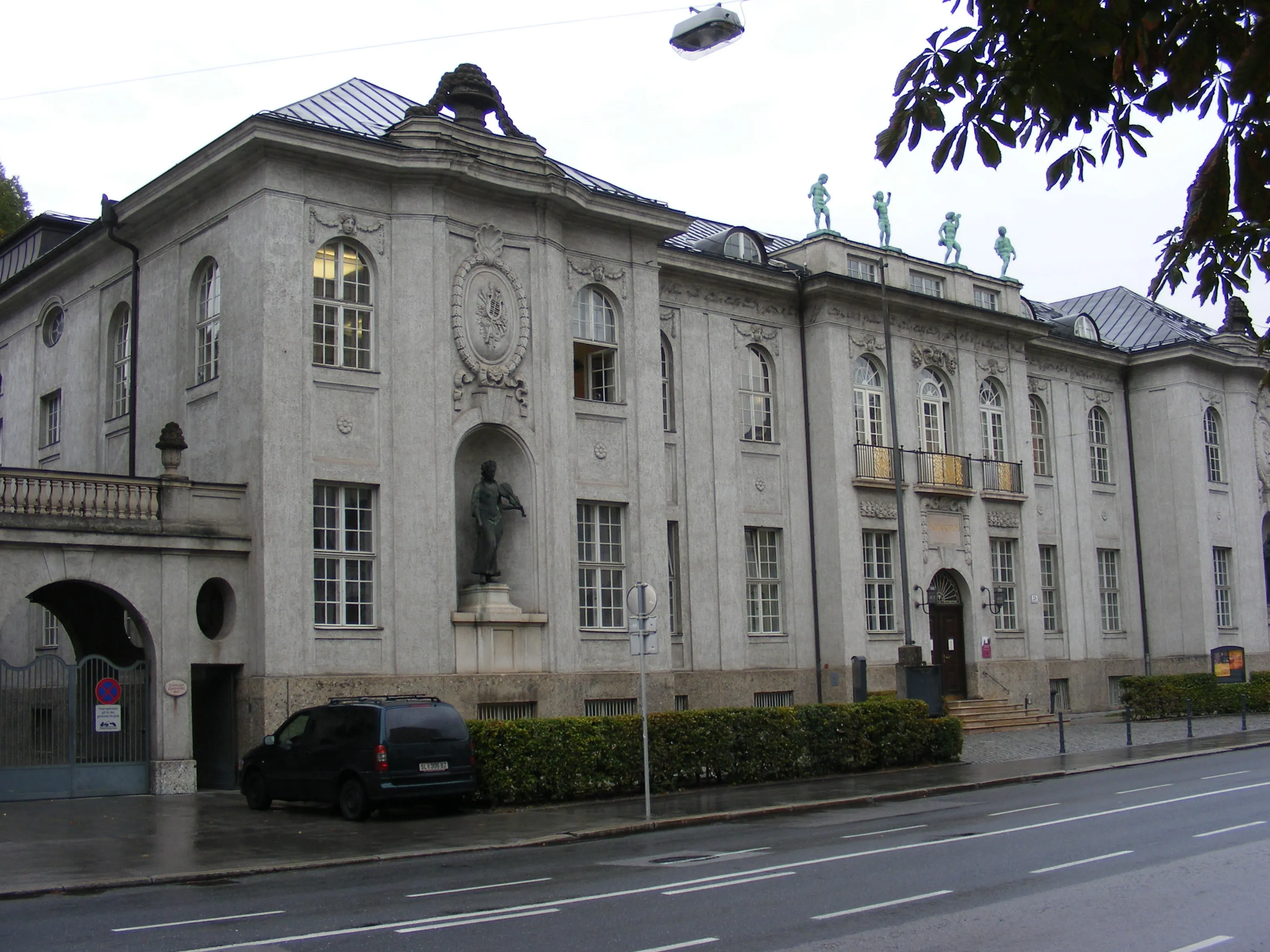Exterior view of the International Mozarteum Foundation building, featuring a classical facade with a central statue in a niche and figures on the roofline, seen from across a wet street.