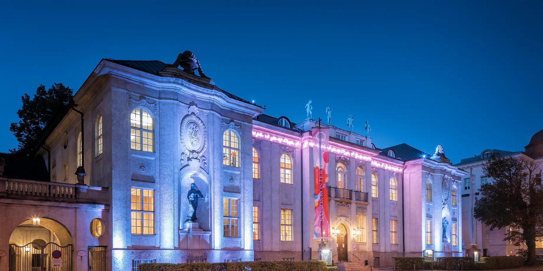 Mozarteum building facade illuminated at dusk with blue and pink lighting, showcasing classical baroque architecture with statues and arched windows