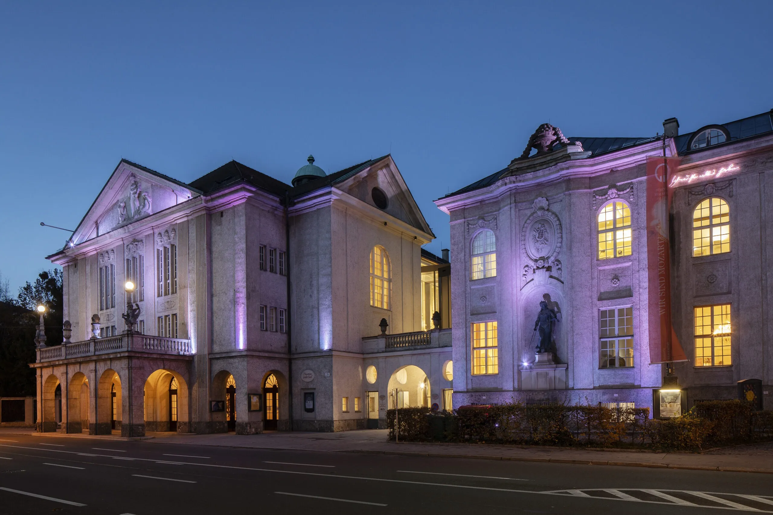Exterior view of the Mozarteum building at dusk illuminated with purple uplighting, showing classical facade with arched entrances and warm interior lights glowing from large windows
