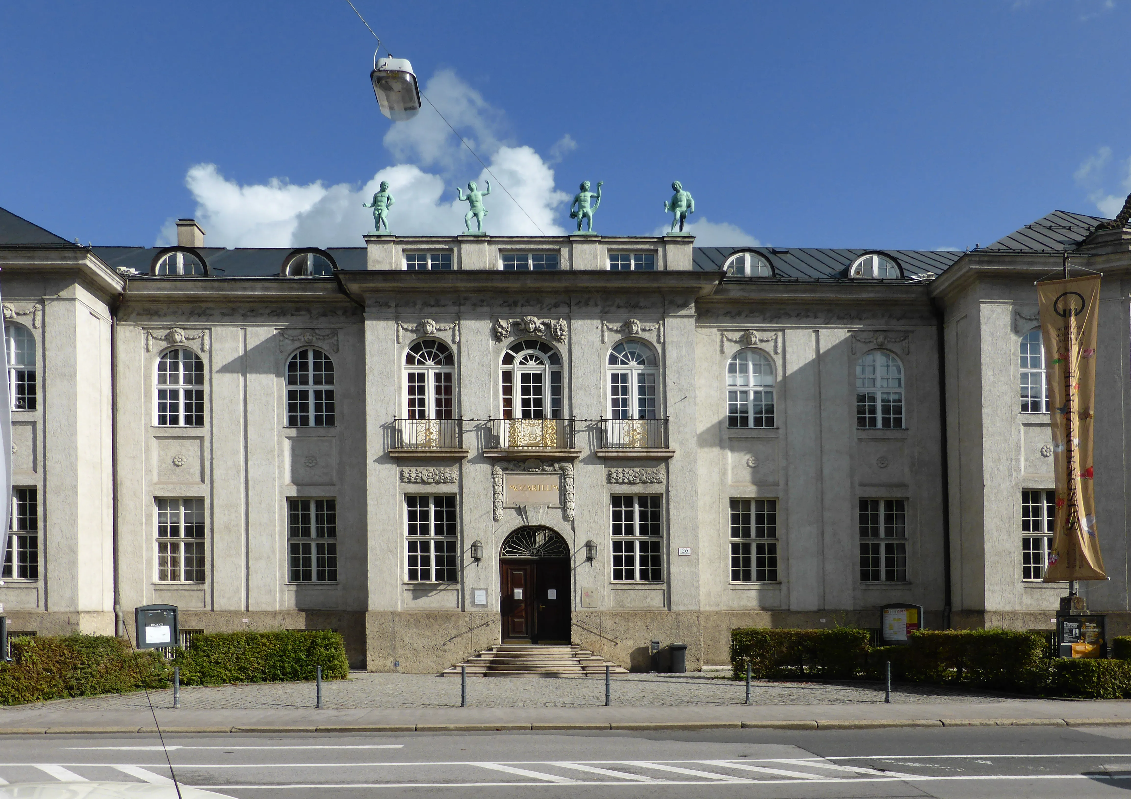 Classical gray stone building facade with arched central entrance, symmetrical windows, four green statues on the roof, and a vertical banner on the right side under a blue sky