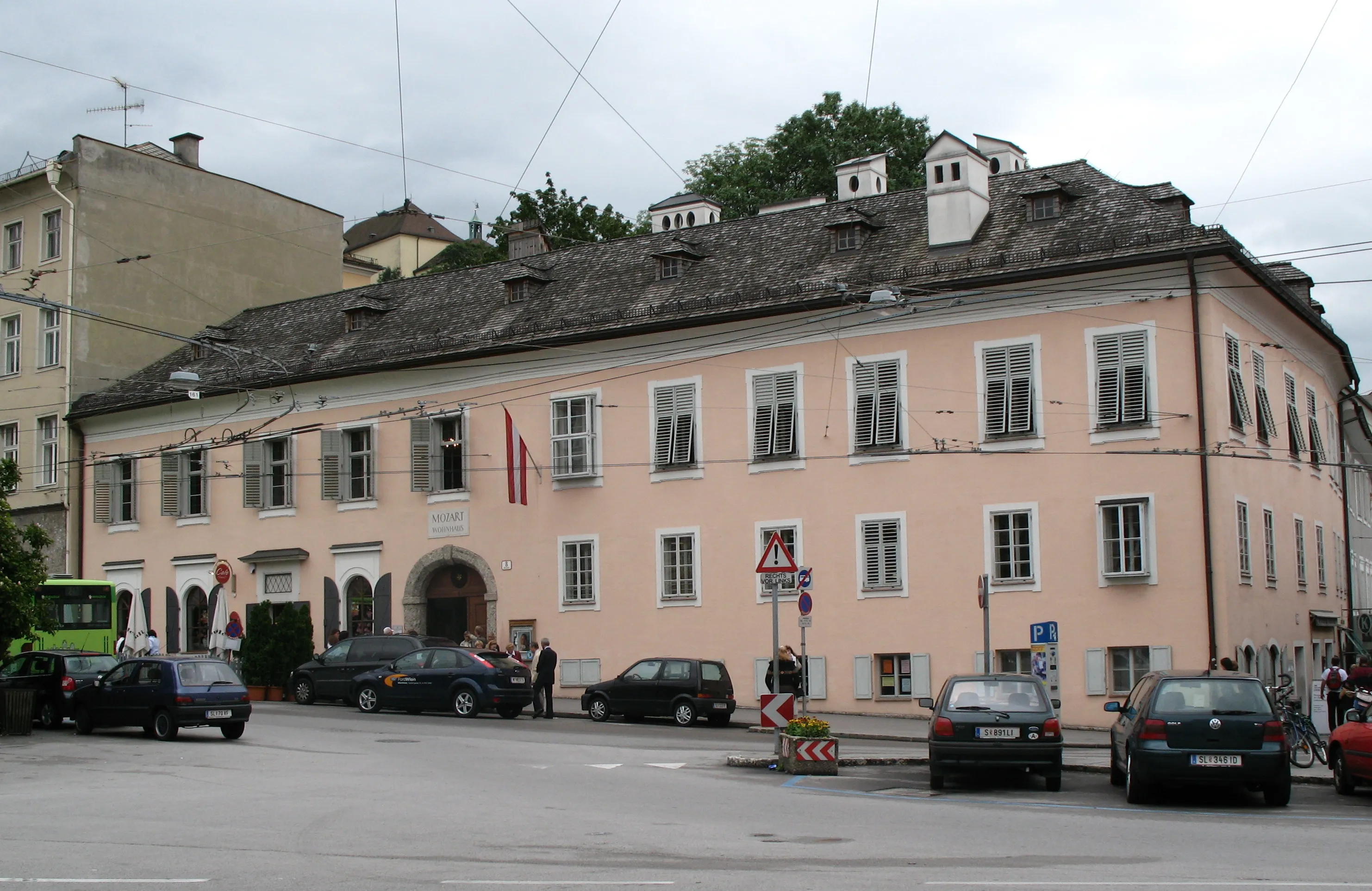 Exterior view of a large, light pink historic building with a dark tiled roof and arched entrance, identified as the Mozart Residence in Salzburg, with cars parked along the street.