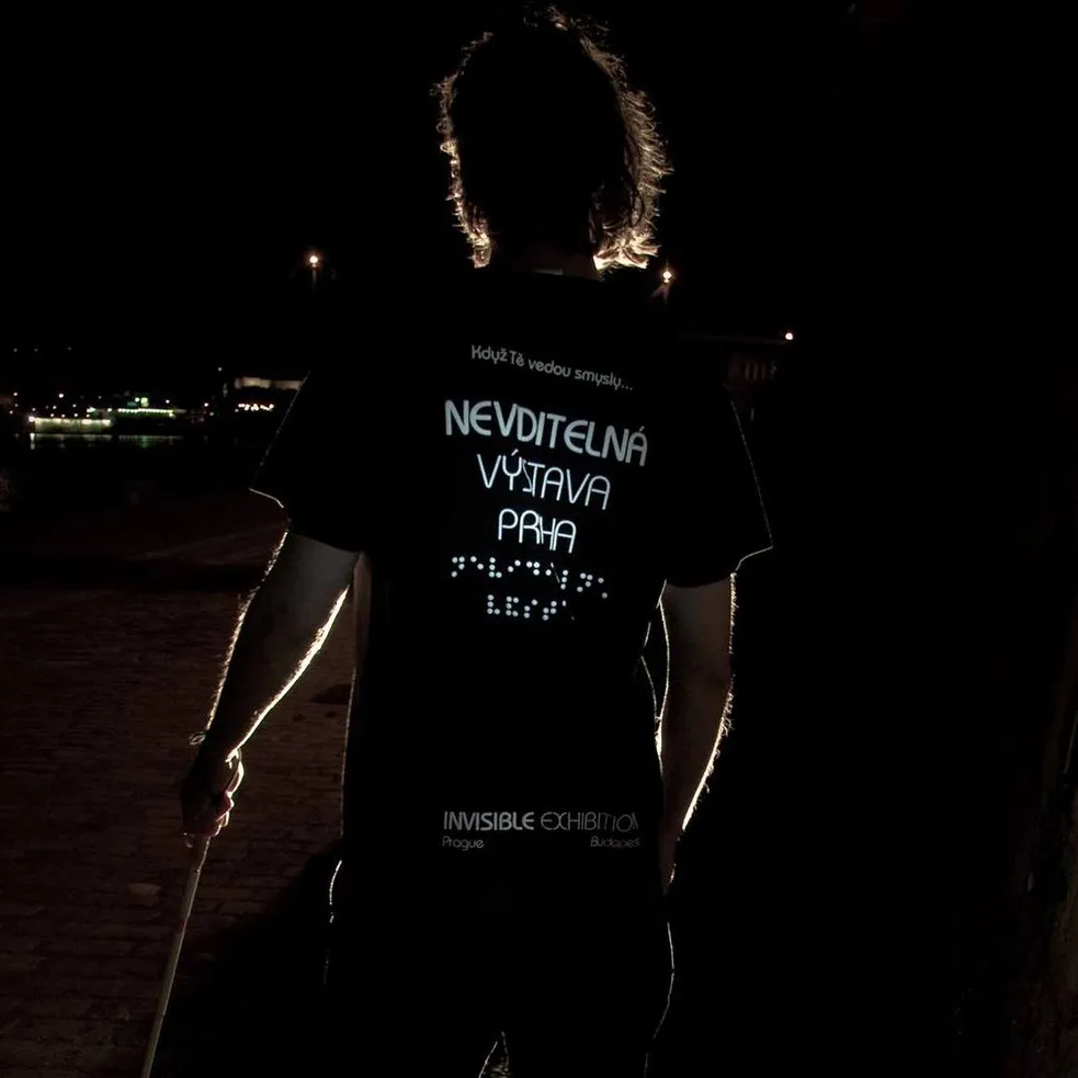 persoon in donkere omgeving met een T-shirt met de tekst 'NEVĎITELNÁ VÝSTAVA PRAHA' en braille