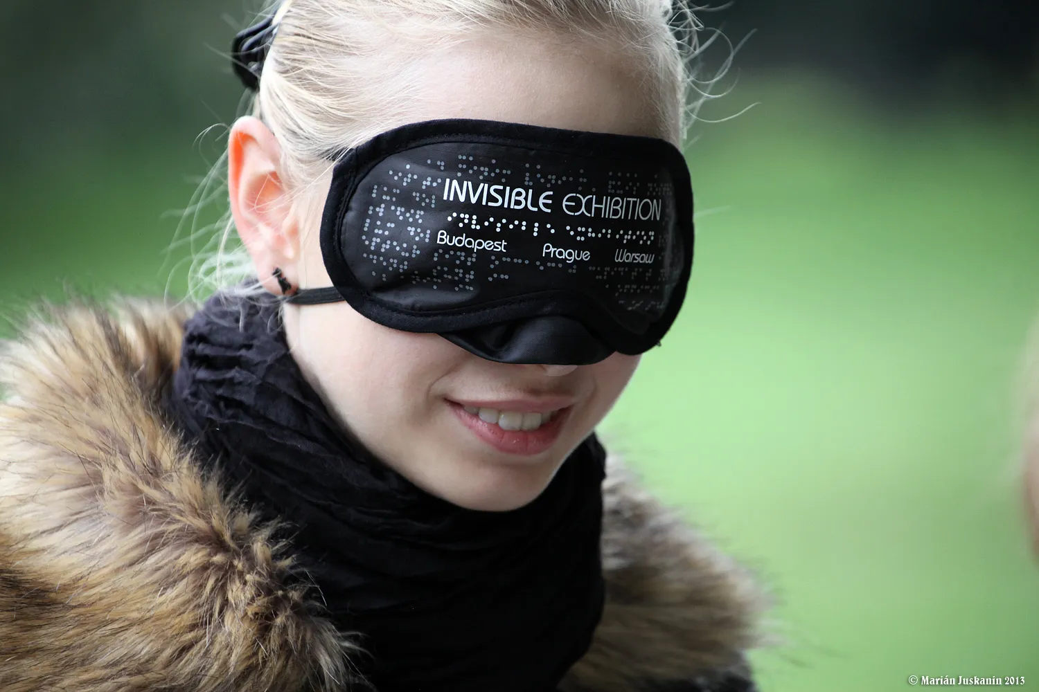 Een persoon met een zwart blinddoek met de tekst 'INVISIBLE EXHIBITION', staand in de buitenlucht met groen op de achtergrond.