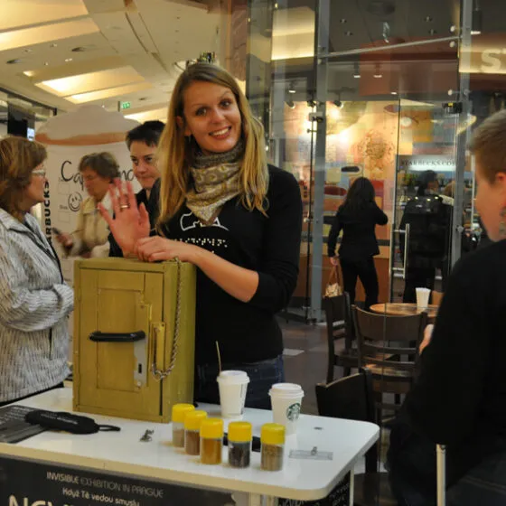 Vrouw glimlacht en zwaait achter een tafel met een gele doos en kleine potjes in een openbare ruimte