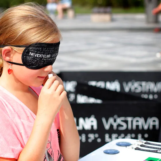 Een jong meisje met een zwart blinddoek met witte tekst en een roze shirt, staand voor een zwarte banner met witte tekst waarop 'NEVIDITELNÁ VÝSTAVA' staat.