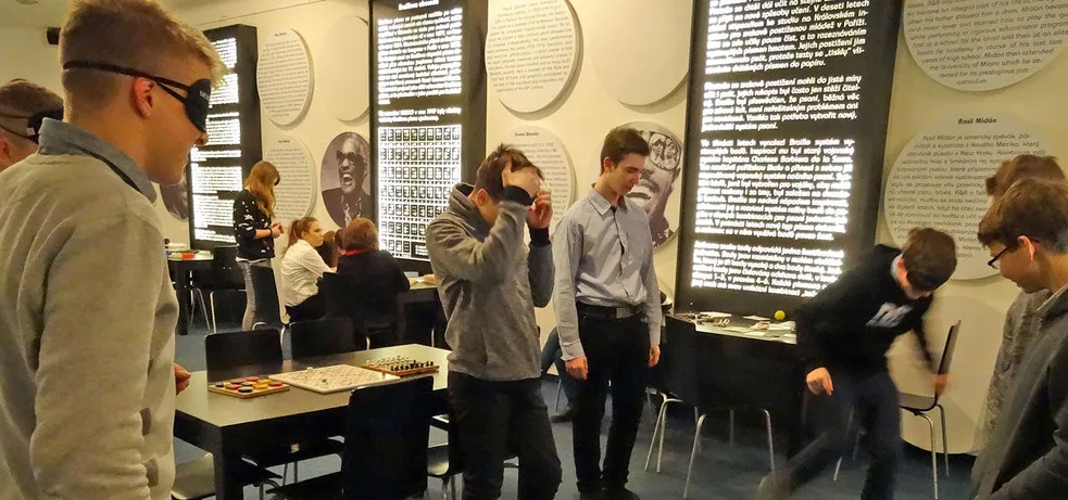 Mensen in een donkere kamer met blinddoeken en anderen die toekijken, omringd door verlichte tekstpanelen aan de muren.