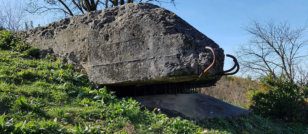 Large concrete bunker fortification with a rusted metal loop, set on a grassy hillside under a clear blue sky