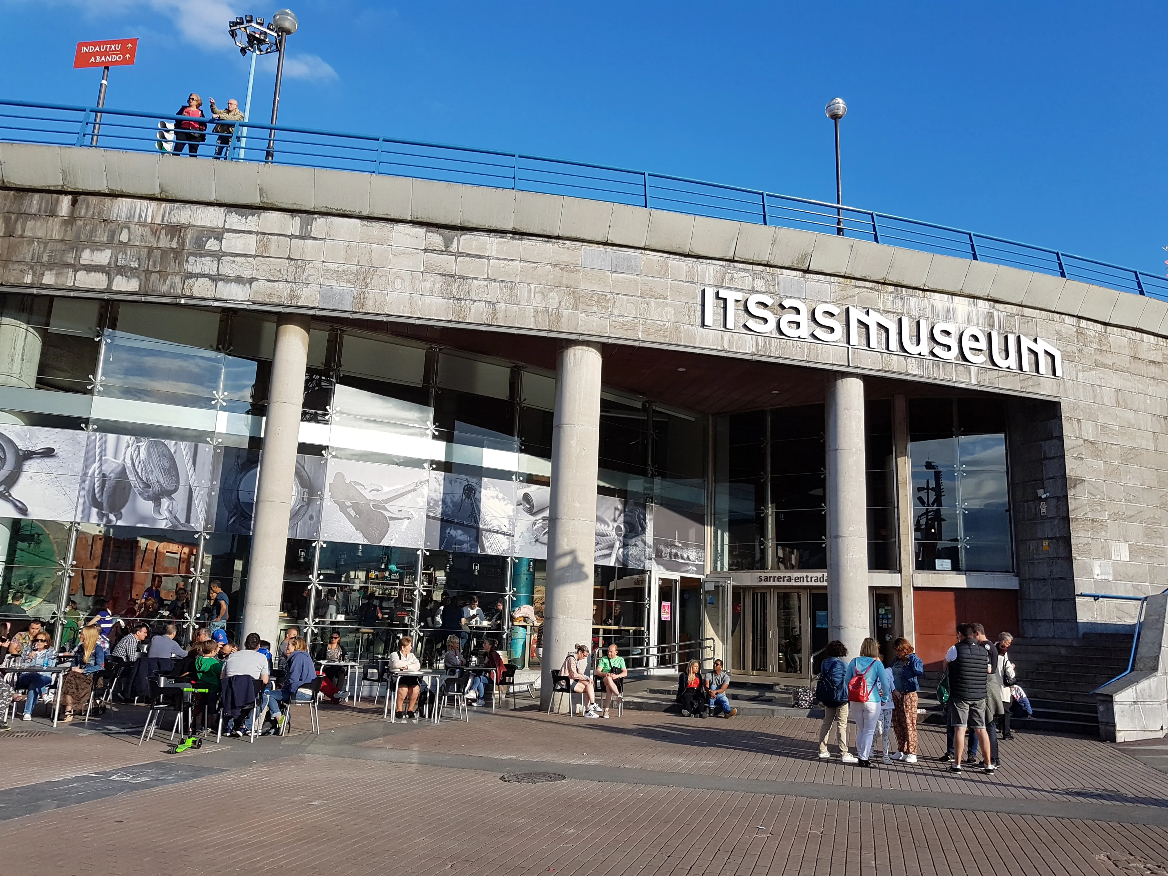 Fachada exterior del edificio del Itsasmuseum Bilbao con su nombre en letras blancas sobre la estructura de hormigón, con visitantes reunidos en la plaza inferior.