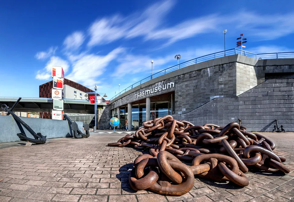 Grandes cadenas de ancla oxidadas amontonadas en una plaza pavimentada frente al edificio del Itsasmuseum Bilbao bajo un cielo azul