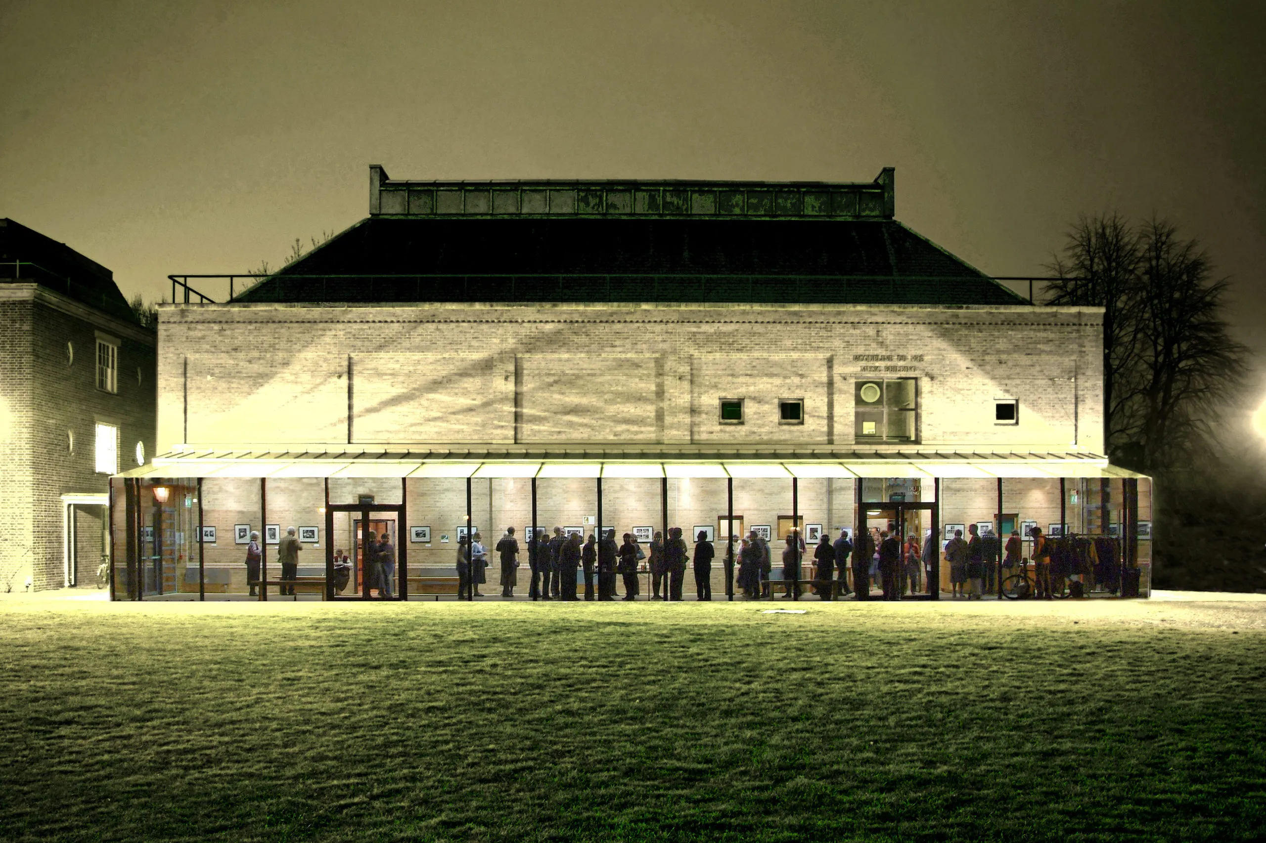 Nocny widok na Jacqueline du Pré Music Building, ukazujący nowoczesną, przeszkloną rozbudowę z ludźmi wewnątrz, na tle oświetlonej, ceglanej sali koncertowej z trawnikiem na pierwszym planie.