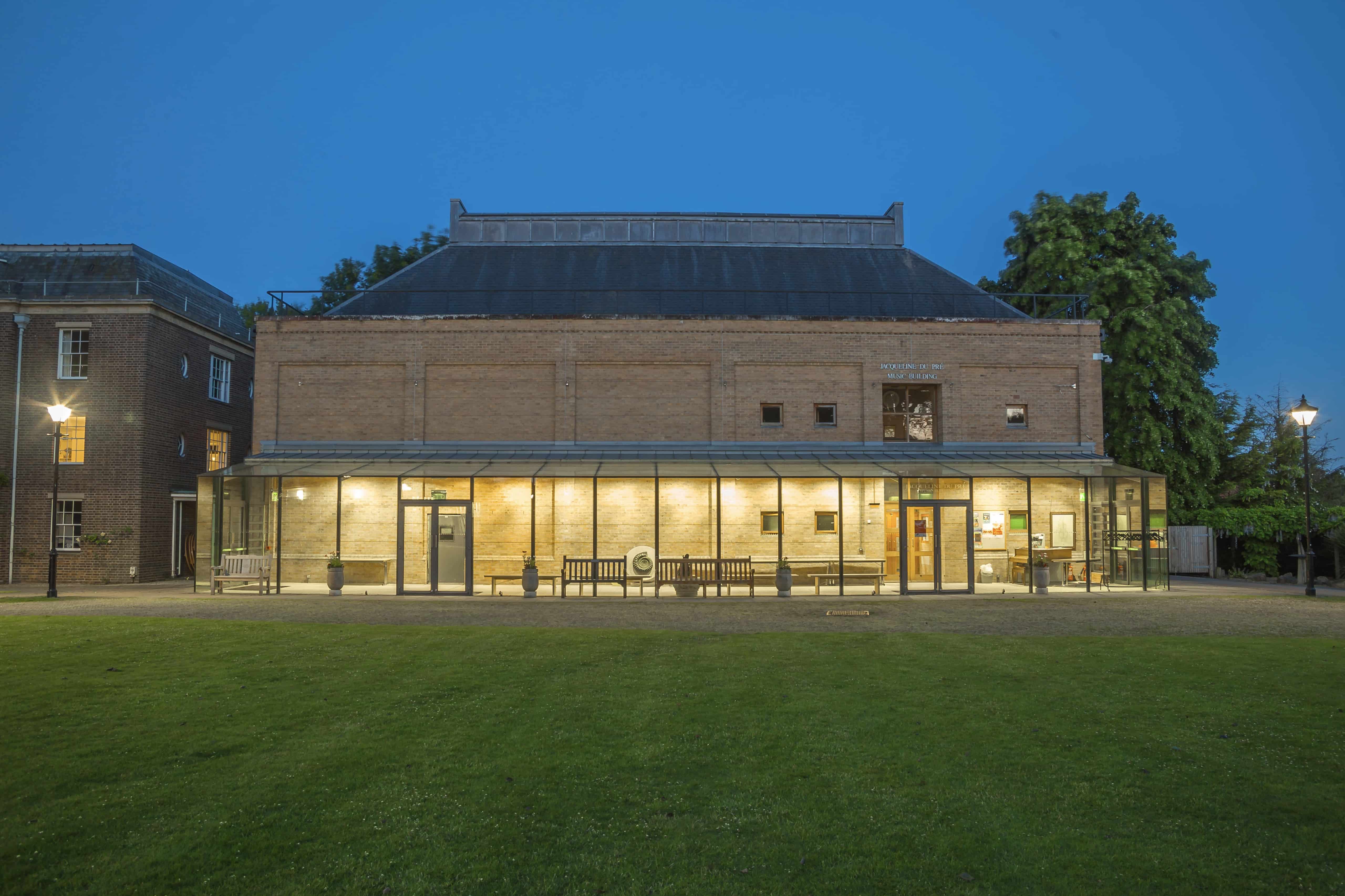 Widok z zewnątrz na Jacqueline du Pré Music Building o zmierzchu, ukazujący oświetlony, przeszklony parter i ceglaną konstrukcję wyższych kondygnacji, z zielonym trawnikiem na pierwszym planie.