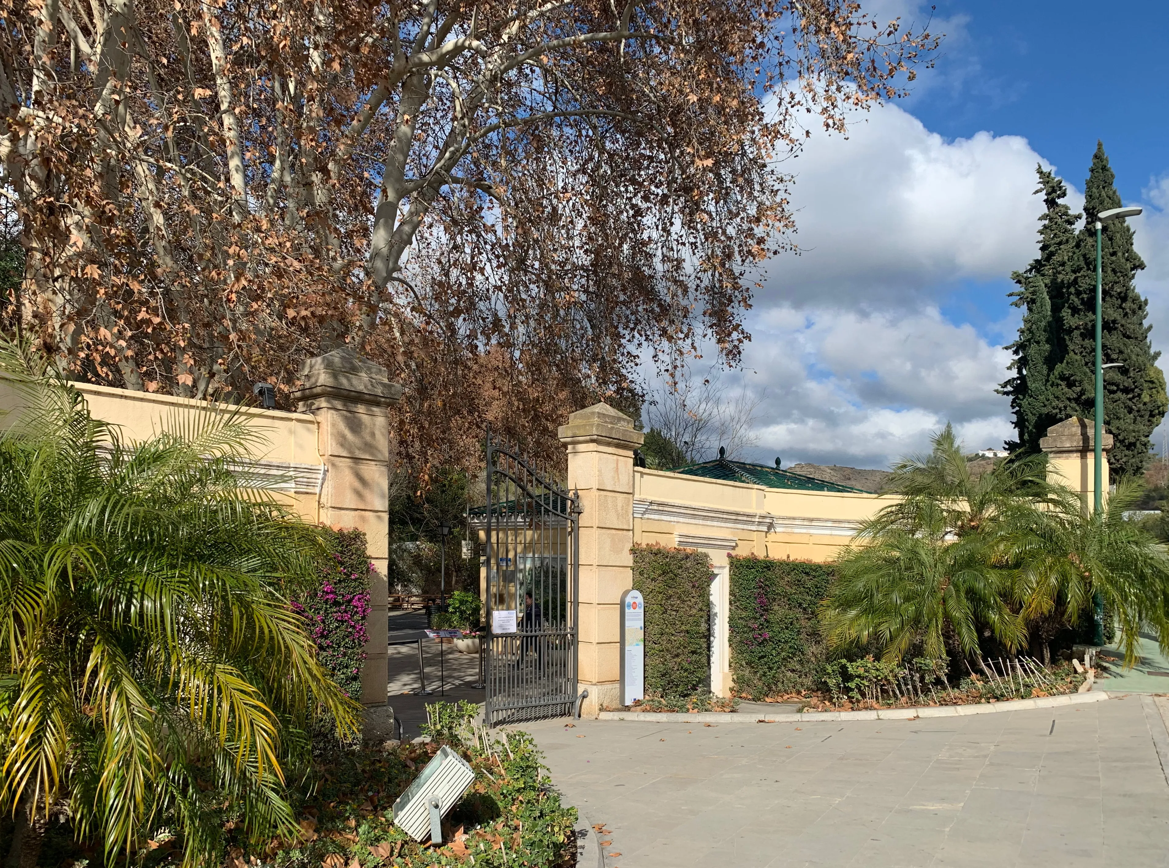 Puerta de entrada de piedra con verja de hierro y muro curvo flanqueado por palmeras en el Jardín Botánico-Histórico La Concepción