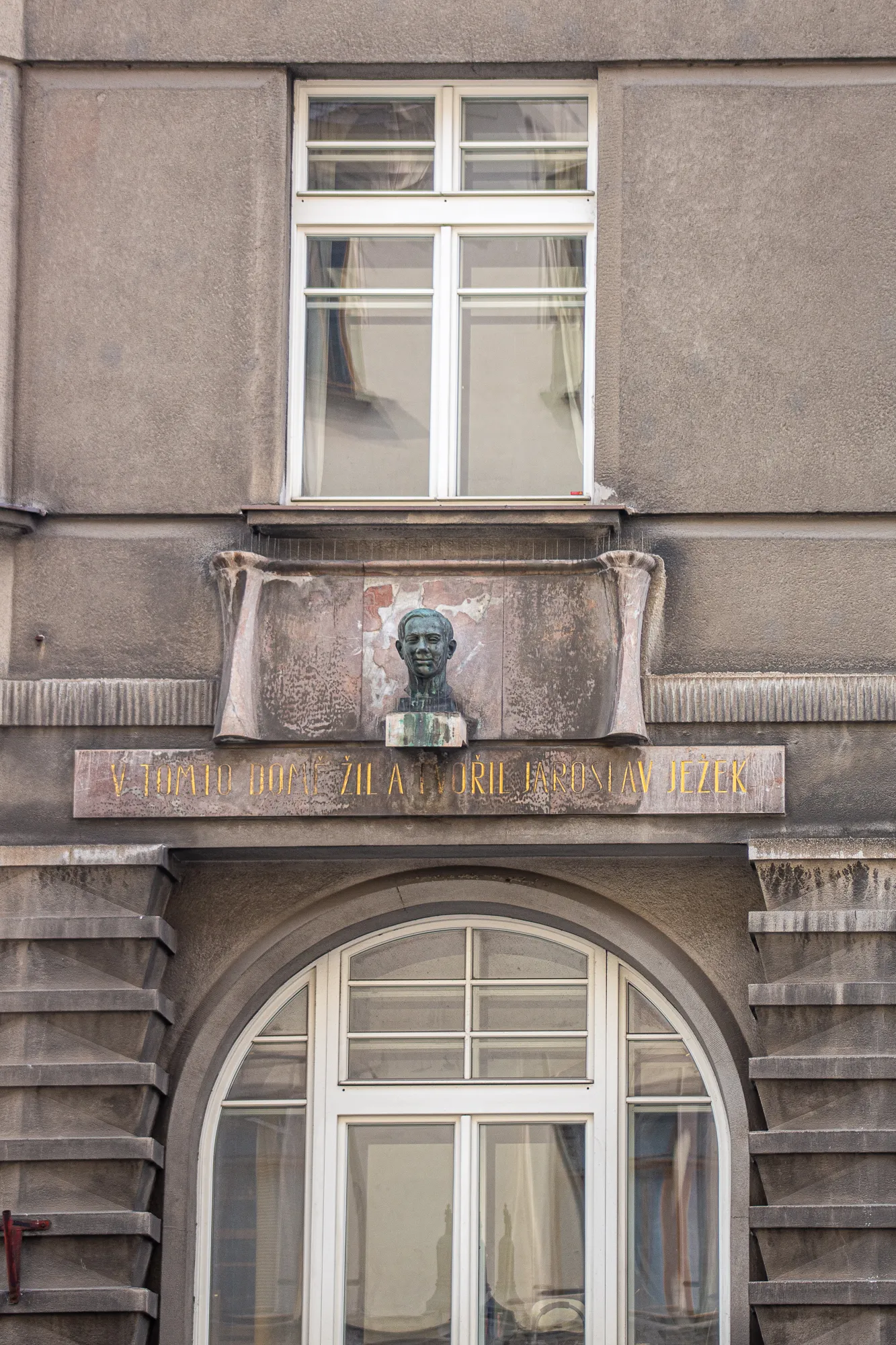 Brązowe popiersie Jaroslava Ježka nad tablicą z jego nazwiskiem, zamontowane na fasadzie budynku z oknem i łukowym portalem poniżej